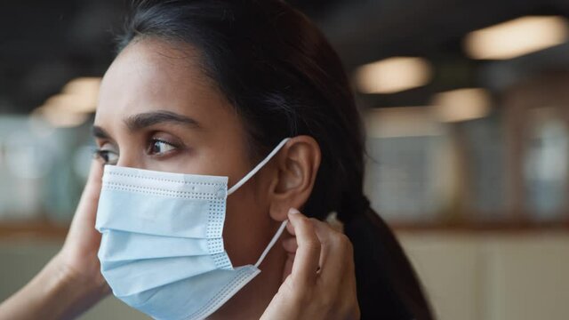 Portrait Of Nurse In Scrubs Putting On Face Mask In Busy Hospital During Health Pandemic