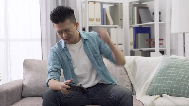front view asian young guy holding wireless controller auto racing at bright home living room, pounding and patting heavily on lap feeling upset because he lost the game
