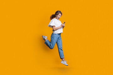 Fototapeta na wymiar Emotional caucasian blonde woman running and jumping on a yellow studio wall while chatting on mobile and promoting something