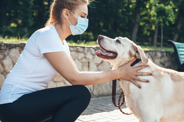 Young blonde woman touching her dog while wear medical mask on face and walk in park