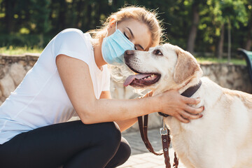 Caucasian woman with medical mask on face is embracing her golden retriever during a walk in park