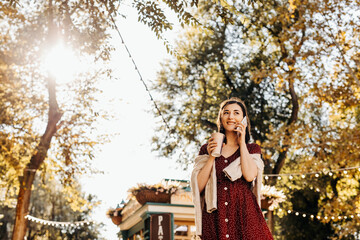 Young woman with a cup of coffee, talking on smartphone, outdoors, on autumn day.