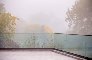 Autumn fog in the old majestic park. Colorful leaves on the trees. Glass panels on the edge of the terrace. Outdoor summer terrace.