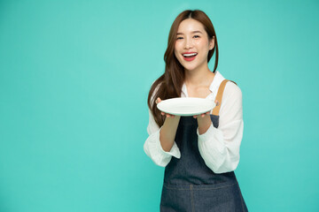 Young Asian woman in apron standing and holding empty white plate or dish isolated on green...