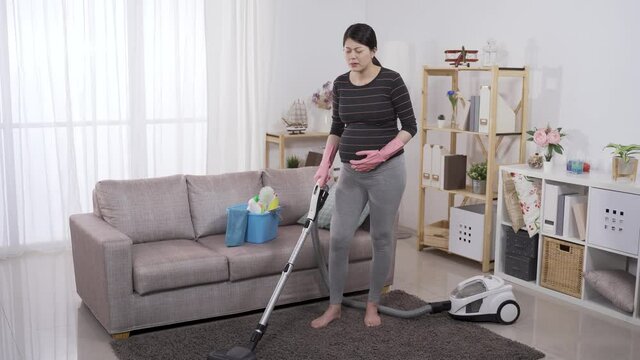 full length asian female in her third trimester feeling sudden painful contraction while cleaning floor with a hoover at home.