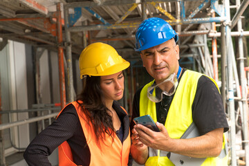 Senior engineer with his young trainee on construction site wearing safety vest, helmet and goggles checking the work plans on a smartphone