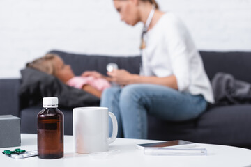 Selective focus of pills, syrup and smartphone on table near mother sitting beside sick child at home