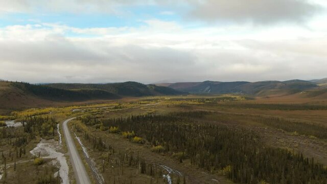 View of Scenic Winding Road surrounded by River, Mountains and Valley in Autumn on a Cloudy Day. Aerial Drone Shot. Taken near Dempster Highway, Yukon, Canada. 4K