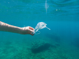 Man collecting a disposable protective face mask thrown in the sea, ocean pollution. Infectious waste from coronavirus