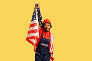 Patriotic afro-american worker in uniform wit dreadlocks holding flag of united states of America, human rights. Indoor studio shot isolated on yellow background