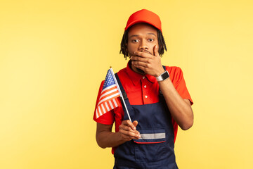 Scared afro-american handyman in uniform with dreadlocks holding usa flag closing mouth with hand,...