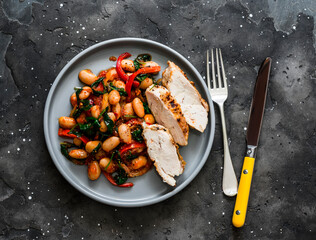 Roasted chicken breast with beans, spinach, paprika, tomato sauce on a dark background, top view....