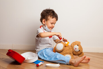 funny curly kid in a medical mask and glasses with a stethoscope on his neck plays a doctor with a cat. The child wants to inject the cat with a toy syringe.