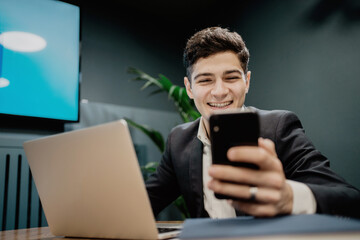 Stylish business suit, fashionable barbershop hairstyle, talking and smiling. a dark-haired man is sitting next to a laptop and holding a phone. office space for work