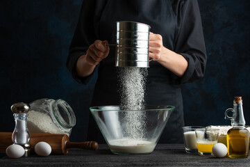 Close-up view of chef in black apron sifts flour into the glass bowl for preparing dough. Frozen motion. Dark background.