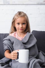 Selective focus of kid wrapped in blanket looking at camera while holding cup on couch