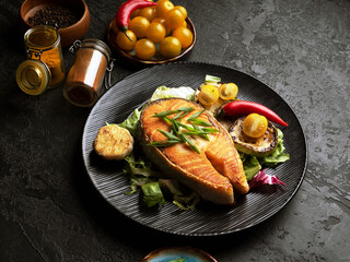 Healthy and diet food concept. Grilled salmon steak and vegetables on the grill on black background. Closeup. Red fish with garlic, yellow tomato and spicies on black plate. Soft focus
