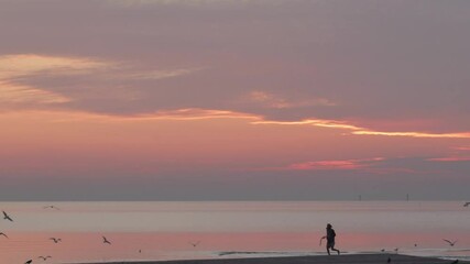 A young man with long hair dressed in a blouse running across the beach in the morning hour during the sunrise. Purple and violet morning sunrise scene with a person.