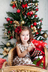 Cute girl in dress sitting on snow wicker sleigh on traditional Christmas tree and gifts background. New year decorations.
