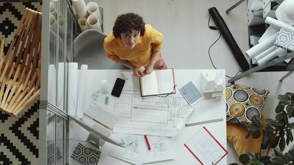 Top down shot of young female architect drawing construction plan on paper, then looking up and smiling at camera while working in office