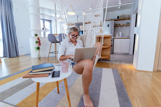 Beautiful young businesswoman working on a laptop computer from home, sitting in a hanging hammock chair in a modern apartment. Lifestyle, modern interior design and work from home concept.