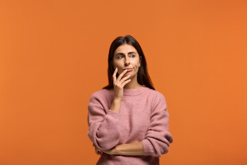 What should i do. Portrait of beautiful woman in pink sweater having doubtful expression, looking away in indecisiveness, holding her finger on a lips, trying to find solution - obrazy, fototapety, plakaty