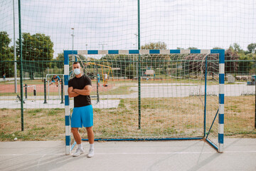 Full length of a young fit male  caucasian athlete with a face mask standing next to a goal on the football field. COVID - 19 coronavirus protection