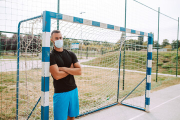 A portrait of a young muscular caucasian male athlete with a face mask standing next to a goal on the football field. COVID - 19 coronavirus protection