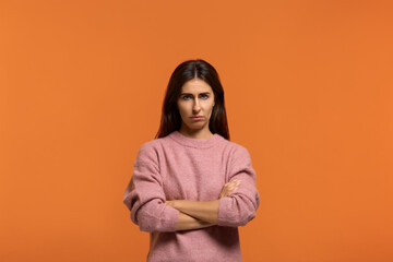 Grumpy irritated looks unhappy. Portrait of serious woman in pink sweater, has serious confident expression, keeps arms crossed, feels angry after quarrel with somebody. on orange wall