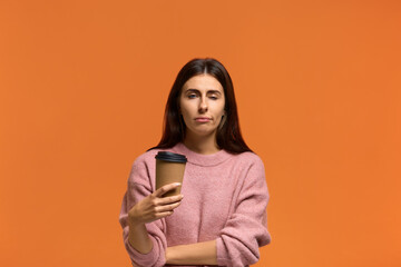 Fototapeta premium Its morning already. Displeased sleepy overworked woman in pink sweater closes eyes from weariness, holding takeaway drink, feels fatigue after learning or working. on orange background