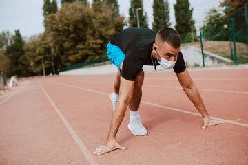 Obraz premium Fit young male caucasian athlete with face mask starting his sprint on an all-weather running track. Muscular athlete outdoors. COVID - 19 coronavirus protection