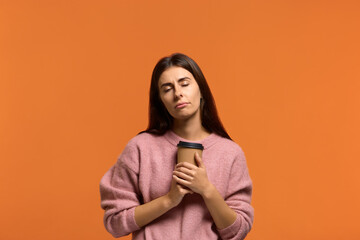 Obraz premium Need to cheer up. Tired sleepy beautiful woman in pink sweater with a tortured look has sad expression, closes eyes, keeps disposable cup of drink containing caffeine. on orange background