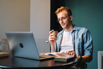 Caucasian man college student in glasses drinking coffee while studying with books laptop distantly preparing for test exam writing essay doing homework at home, distantly education concept.