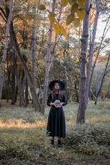 Young beautiful cute woman in dark dress and witch's hat holds a skull in her hands. Halloween party costume. Forest, park with autumn trees.