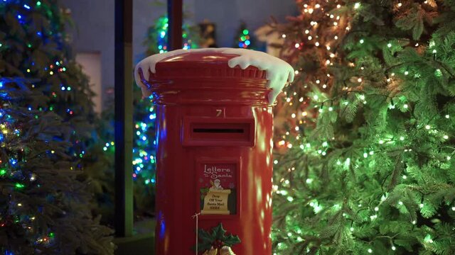 Beautiful decorated red mailbox of Santa Claus standing among christmas trees with glowing garlands, special winter box for putting hopeful letters and greeting postcards to Santa in lapland