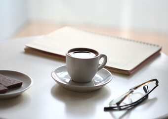 A cup of coffee on a white table, next to chocolate, a notebook and glasses. Behind the window