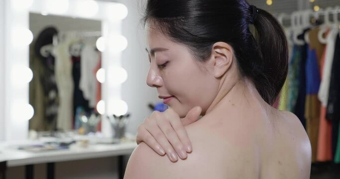 slow motion of close up smiling elegant woman actress cares about her shoulder applying cosmetic cream. half naked young asian girl in backstage dressing room doing skin protection treatment on back.