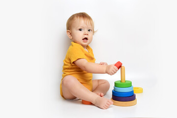 little cute baby under one year old in a yellow bodysuit sits on the floor and plays with a rainbow multicolored pyramid on a white background, place for text