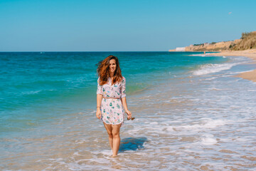 Fototapeta premium Beautiful smiling woman in a dress walking on the sand along the emerald sea, leisure and tourism concept
