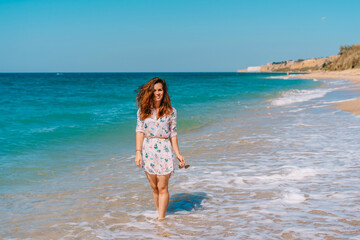 Beautiful smiling woman in a dress walking on the sand along the emerald sea, leisure and tourism concept