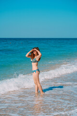 Fototapeta na wymiar A young woman in a bright swimsuit and long hair walks on the sand along the sea with waves, clear azure water