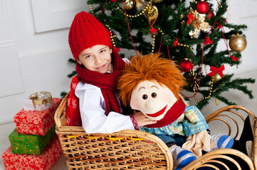 Cute boy in red knitted hat and scarf sitting on snow wicker sleigh on traditional Christmas tree and gifts background. New year decorations.