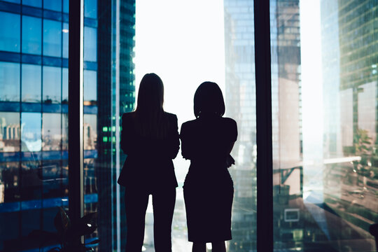 Back view of female colleagues in formal wear standing near window looking at modern exterior of skyscrapers in business center, silhouette of women together planning future success of brainstorming