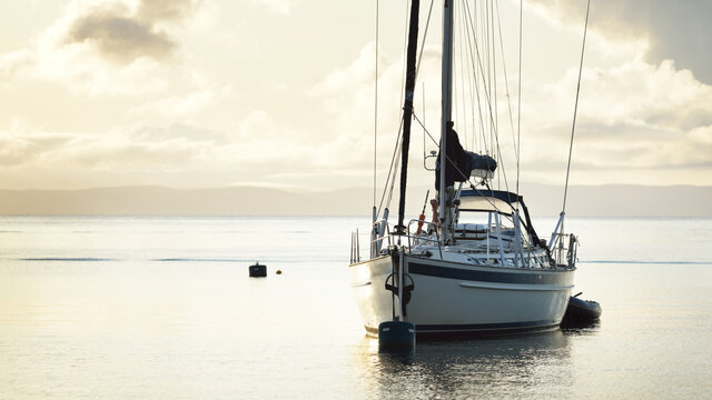 A two-masted sailboat (ketch) with a motorboat anchored on mooring near the shore of Craighouse at sunset. Golden evening light, dramatic cloudscape. Jura island, Inner Hebrides, Scotland, UK
