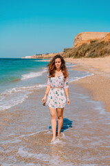 A young woman in a dress and long hair walks along the coast by the sea with waves, the concept of a relaxing holiday