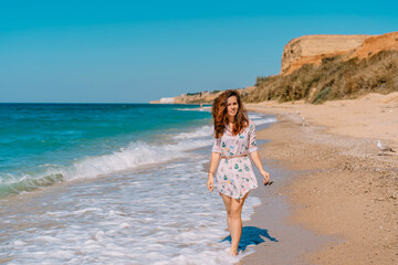 Fototapeta premium A young woman in a dress and long hair walks along the coast by the sea with waves, the concept of a relaxing holiday