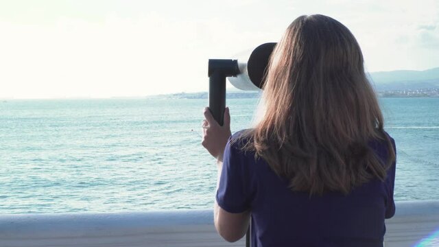 Young woman looks through binoculars on embankment. Media. Back view of attractive woman walking to binoculars on background sea horizon on Sunny day. Observation deck on sea