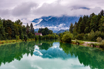 green river on the edge of the forest with a house on the background of a mountain shrouded in fog