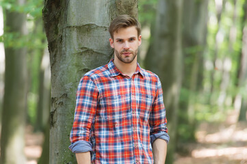 perfect hair. casual fashion style. muscular man in forest. sexy macho in unbuttoned shirt outdoor. young unshaven man has groomed hair. male beauty and fashion. guy with bristle wear checkered shirt