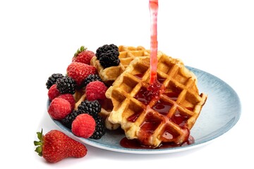 a blue plate of Belgian waffles with strawberries, raspberries and blackberries strawberry syrup being poured isolated on white